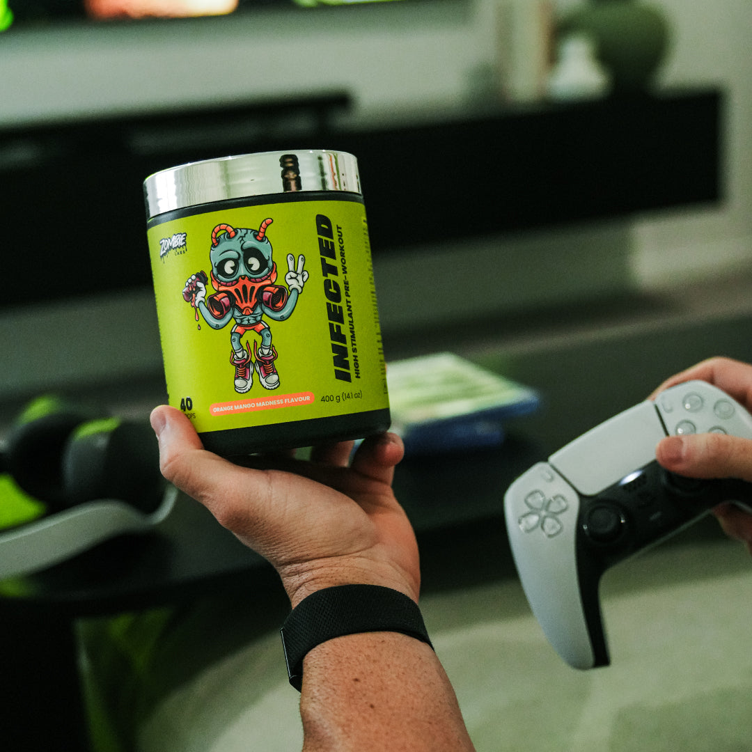 Person's hand holding Zombie Labs Infected Preworkout in Orange Mango Madness flavour and a game control