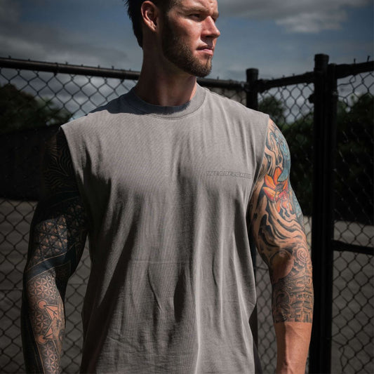 A muscular man with tattooed arms wearing a grey sleeveless tank top from Zombie Labs, standing outdoors next to a chain-link fence under a cloudy sky