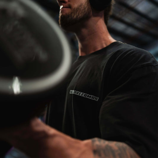 Close-up of a man lifting weights in a gym, wearing a black 'Pump Cover' tee from Zombie Labs. The shirt has the 'WEAREZOMBIE' logo on the chest.